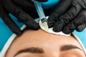 Concept of plastic surgery. Close-up of female's head getting injection in the cosmetology salon. Cosmetologist in latex gloves with syringe injects a botulinum toxin in forehead