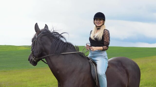 Glamorous young woman in a helmet, trendy blouse with translucent lush sleeves and light jeans sitting on a black horse