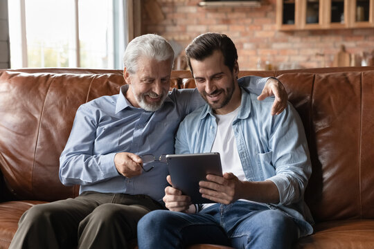 Happy grown son teaching senior father to use tablet. Two family generations men with digital device sitting on couch together, browsing internet, reviewing family pictures, making video call - Powered by Adobe