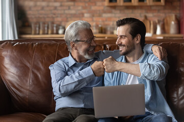 Happy excited grown son and elderly 70s father with laptop sharing good news, celebrating success, giving fist bumps. Two male generation of fans watching online sport game. Fatherhood, family leisure