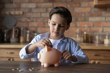 Focused school kid saving money for purchase, putting cash in pink piggy bank at home. Preschooler...