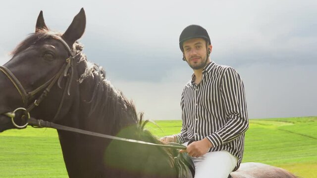 A young man in a helmet, striped black and white shirt and light trousers sits on a black horse in the field. A non-professional rider holds the reins