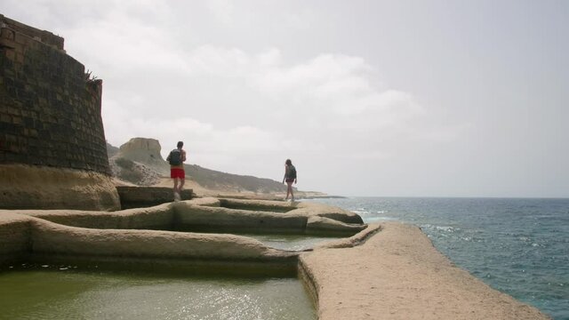 Hikers travel on Salt Pans on the Island of Gozo in Malta. Naturally created geometric water filled salt pans. Rear view hikers travelers with back pack enjoying nature beauty on coast of salt lake.