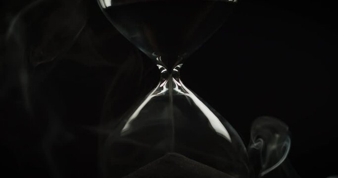Light revealing an hourglass with black sand and a smoke wrapping it on a black background. 