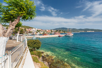 Seascape featuring a pier in front of a Mediterranean town with small boats for tourist trips with clear water near the coast and light clouds on the blue sky