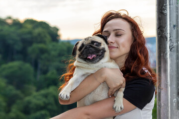 Young red-haired woman hugs her pug dog in the park.
