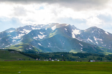 A view of a huge mountain range at the foot of which are houses. Snow can be seen on the mountain and there is beautiful green grass at the foot.