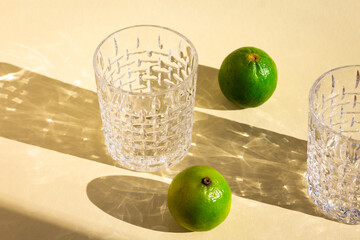 a line of empty crystal glasses and two limes under hard sunlight, glitter reflection on beige background, long harsh rectangular and oval shadows