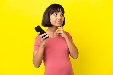 Young mixed race pregnant woman isolated on yellow background using mobile phone and thinking