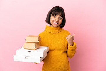 Pregnant woman holding pizzas and burgers isolated on pink background pointing to the side to present a product