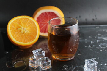 Apple citrus dark juice in a glass next to lies citrus orange grapefruit ice on a black background. Refreshing drinks, summer, soft drinks