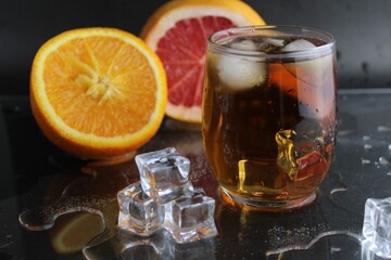 Apple citrus dark juice in a glass next to lies citrus orange grapefruit ice on a black background. Refreshing drinks, summer, soft drinks