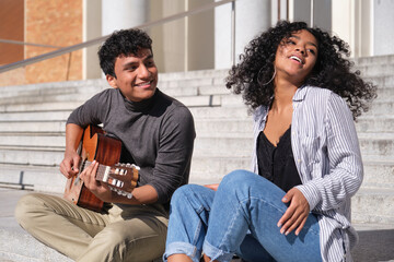 A young latin couple playing the guitar and smiling sitting on stairs. Millennial generation.