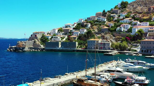 Drone lifting off over small marina in Hydra, Greece. There is a small lovely town on the mountainside, and a boat leaving the harbour. It is a beautiful summer day. Drone aerial 4K