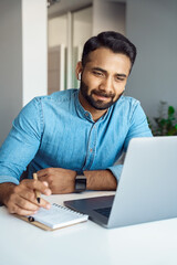 Portrait of handsome young indian man student watching online webinar using laptop and earphone making noes in notebook. Video conference call, business training tutorial, coaching course concept