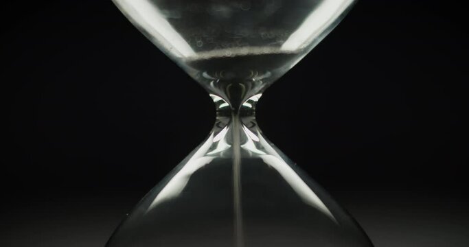 Black sand pouring inside the modern hourglass on a black background. The end of time cycle. Macro shot.