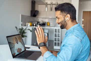 Mature eastern Indian man sitting at home desk front of laptop greeting business partner on screen....