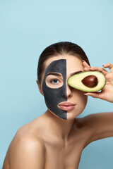 Beauty Facial Mask . Beautiful Young Woman with black mask of clay on face .Skin care .Girl beauty model holding half an avocado in hand . Cosmetic  spa mask . Facial treatment