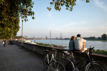 Outdoor sunny view, couple sit and chill out on balustrade at Rheinuferpromenade along riverside of Rhine river in Düsseldorf, Germany. - obrazy, fototapety, plakaty