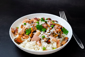 Seafood risotto in white plate on black background. Rice with mussels, baby octopus, squid, shrimps and parsley stewed in white wine. Traditional Spanish seafood paella. Healthy Mediterranean cuisine.