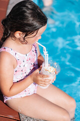 top view of a child in a pink swimsuit drinking a cocktail, sitting on the edge of the pool in a family hotel on vacation. 