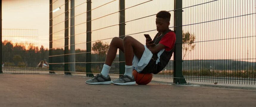 African American teenager sits on a ball and using his phone on basketball court. Shot with 2x anamorphic lens