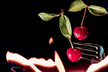 Two red cherries stuffed on a metal fork. Cherries with two green leaves on a black background. Orange sparks and flames float all around. Abstraction.