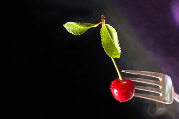 Red cherry stuffed on a metal fork. Cherry with two green leaves on a black background. White smoke is hovering all around. Abstraction.