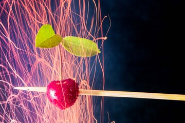 Red cherry stuffed on a wooden toothpick. Cherry with two green leaves on a black background. Sparks and purple, navy blue smoke float all around. Abstraction.