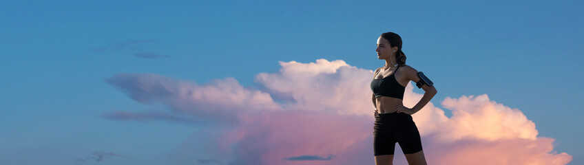 A sporty slim girl in leggings and a top is standing near a concrete wall with a training mat, resting between exercises.