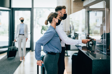 Young couple with protective face masks at the reception of a hotel checking in. Business trip, Coronavirus, Covid-19, safe travel concept.