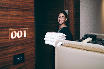 Beautiful young hotel chambermaid in uniform bringing clean towels and other supplies to hotel room.