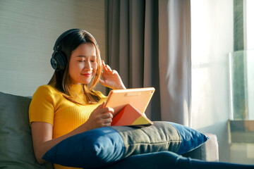 Asian woman sit on sofa listening to the music from headphones while working on digital tablet for online business project at apartment. Female freelance work job plan or send online message at home.