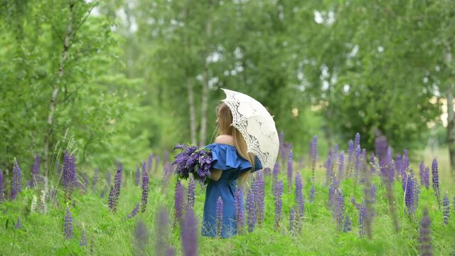Portrait of a young blonde woman with an umbrella in a blue dress with a bouquet in the park. Attractive young girl in the flowered meadow in the spring time. Healthy Lifestyle Concept.