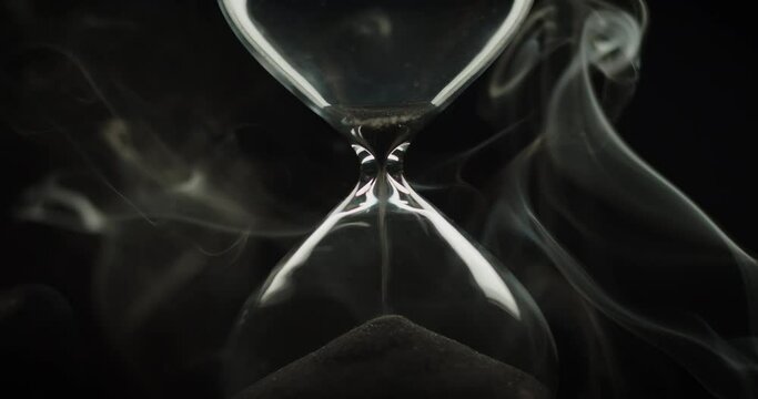 Beautiful smoke surrounding an hourglass with black sand pouring inside of it. Abstract time concept. End of time cycle. 