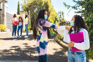 Selective focus of two masked Latina teenage female students with social distancing elbow greetings with another group of out-of-focus female classmates in the background.