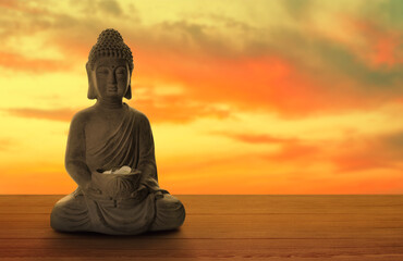Beautiful stone Buddha sculpture with flower petals on wooden surface at sunset. Space for text