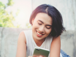 Attractive Asian female with black short hair in casual white sleeveless shirt smiling while using smartphone on building and green nature background.