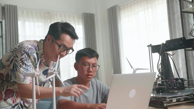 Cheerful Father And His Son Using Laptop Computer Programming Electric Wind Turbine Model And 3D Printer At Home
