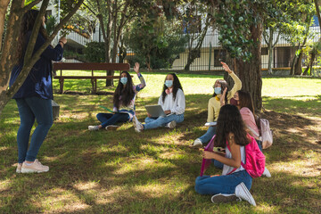 Masked Latina teenage students raise their hands during an outdoor class with a teacher