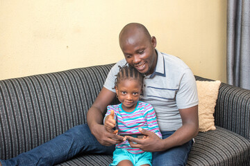 A happy African father or guardian sitting on a sofa together with her little daughter that is...