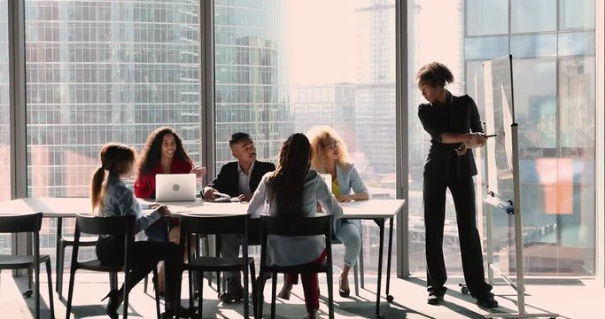 Confident young african american female leader presenting business ideas near whiteboard to concentrated diverse colleagues teammates at brainstorming meeting in modern office with panoramic view.