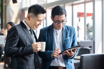 Two Asian businessmen wearing suit discussing and using digital tablet with happy and smiling while...