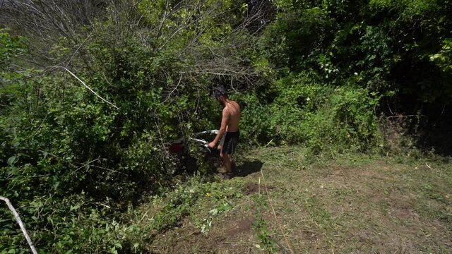 A shirtless man trimming brambles bush and bad weed with flail mower at the backyard - high angle shot