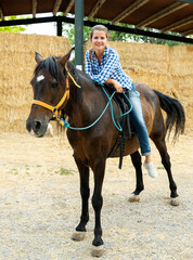 Portrait of happy young woman in jeans and checkered shirt riding bay horse on farm
