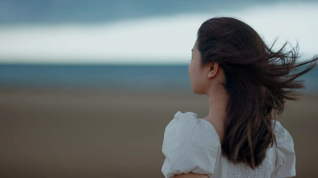 A cute Asian girl is walking on the beach by the sea and the clouds are about to fall and the wind is strong, 4k resolution.