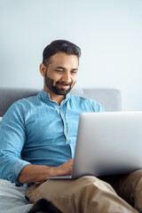 Smiling indian business man having informal video call chat on laptop sit on bed