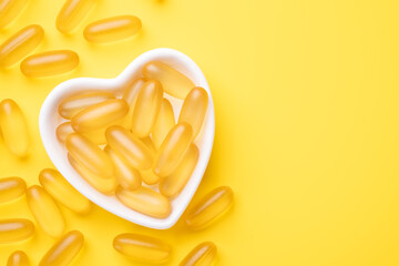 Vitamin D and Omega 3 fish oil capsules supplement in a heart-shaped plate on yellow background....