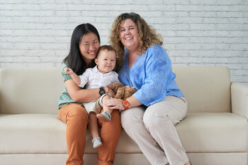 Joyful young LGBT family with little baby boy sitting on sofa at home and looking at camera