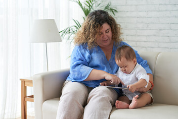 Woman showing educational video on digital tablet to her curious little son sitting on sofa